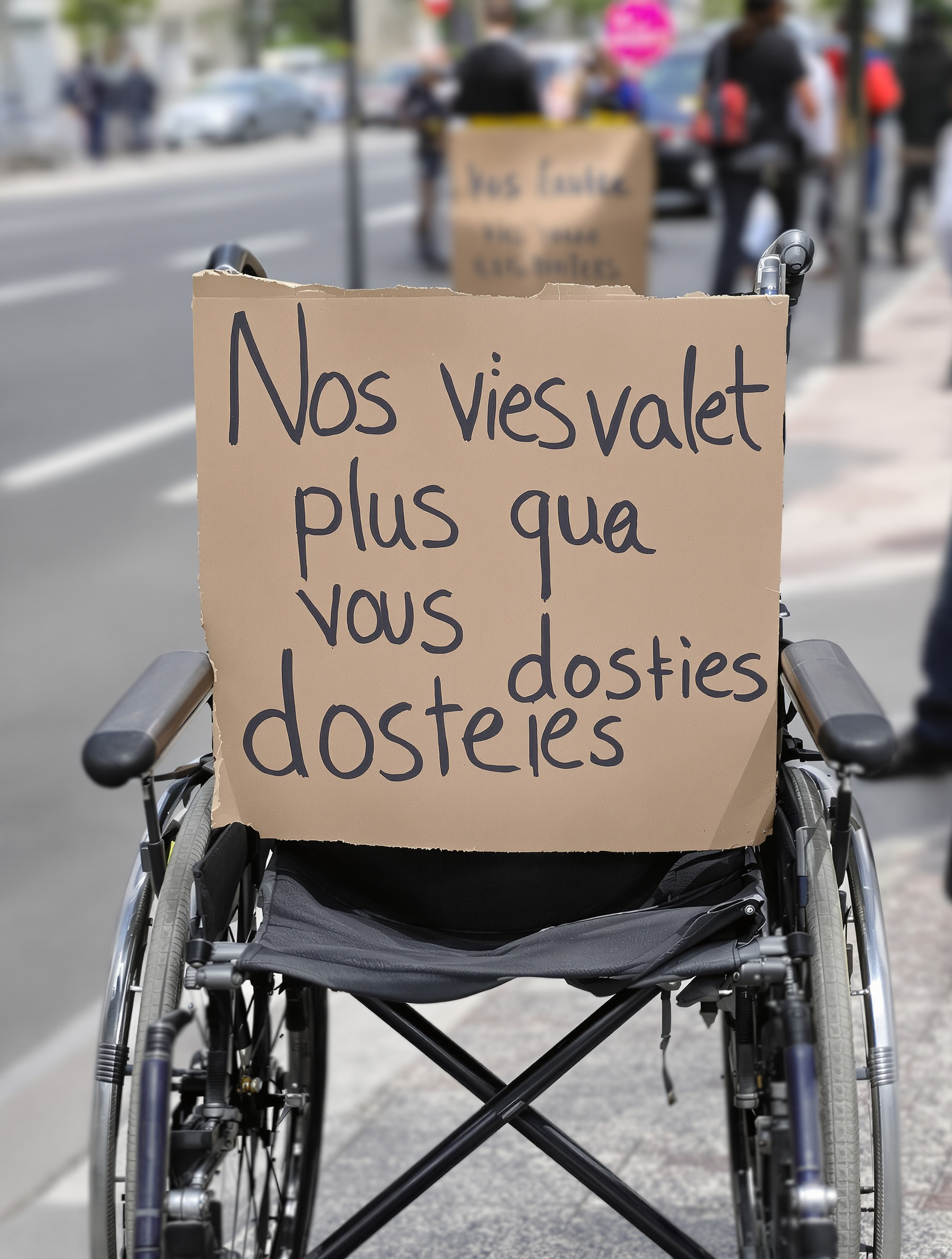 Pancarte sur fauteuil roulant vide : 'Nos vies valent plus que vos dossiers'
